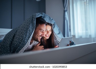 Happy young couple lying in bed at night , holding a tablet