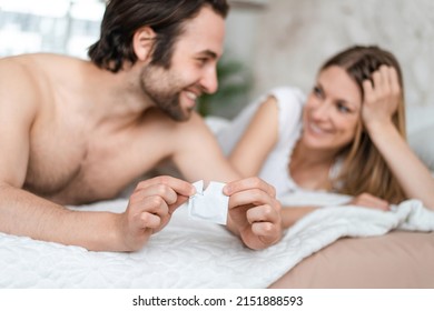 Happy Young Couple Lying In Bed And Opening Condom, Selective Focus. Cheerful Millennial Lovers Choosing Safe Sex, Using Contraception, Copy Space. Pregnancy And STD Control Concept