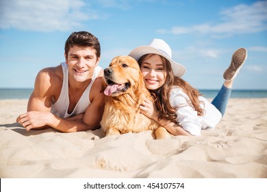 Happy Young Couple Lying And Hugging With Dog On The Beach