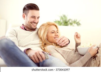 Happy Young Couple Lying On The Sofa At Home With Popcorn Watching TV