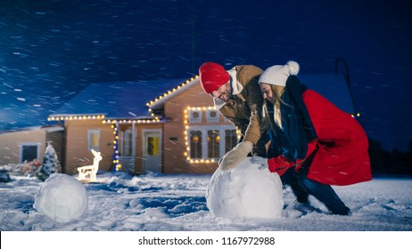 Happy Young Couple Makes A Snowman, Both Rolling Big Snowball. Family Having Fun On One Winter Evening. In The Background House Decorated With Garlands.