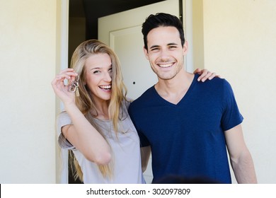 Happy Young Couple Man And Woman Handing Their New Home Keys In Front Of An Open House Door