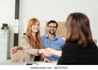 Happy young couple meeting with a broker in her office leaning over the desk to shake hands, view from behind the female agent
