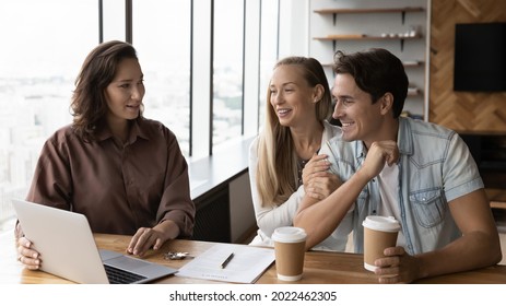 Happy Young Couple Meeting With Real Estate Agent In Office, Filling Agreement, Getting Key, Watching Virtual Apartment Tour On Laptop. Married Clients Consulting Advisor Over Cup Of Coffee