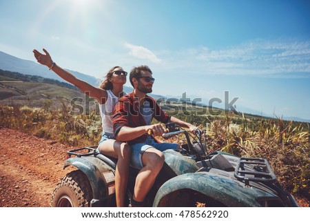 Happy young couple in nature on a quad bike. Young man and woman enjoying a quad bike ride in countryside. Man driving and woman enjoying the ride with her hands raised on a summer day.