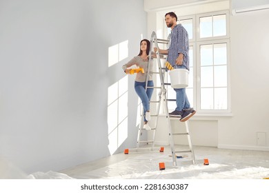 Happy young couple painting the wall of their new home holding paint rollers and standing on the ladder. Married man and woman doing repair renovation preparing to move into a new flat.