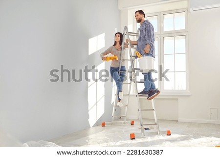 Happy young couple painting the wall of their new home holding paint rollers and standing on the ladder. Married man and woman doing repair renovation preparing to move into a new flat. Foto stock © 