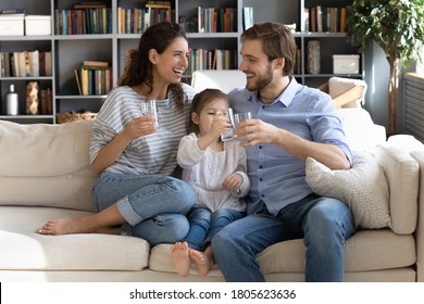 Happy young couple parents teaching little preschool daughter drinking clear water every day. Smiling healthy family holding glasses with pure aqua, enjoying morning daily healthcare habit at home.