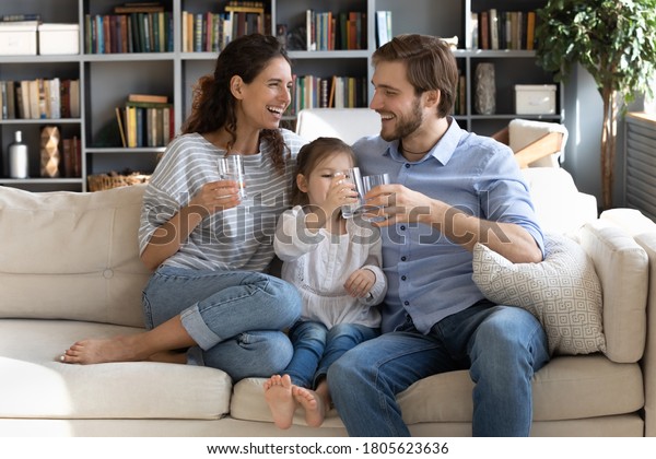 Happy young couple parents teaching little\
preschool daughter drinking clear water every day. Smiling healthy\
family holding glasses with pure aqua, enjoying morning daily\
healthcare habit at home.