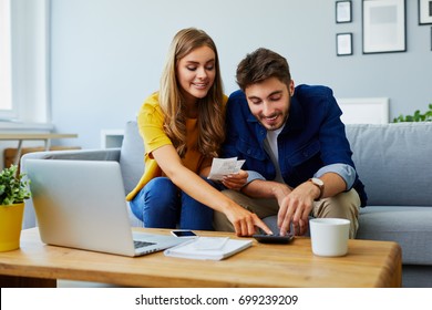 Happy young couple paying bills together and managing budget, sitting on the sofa and using calculator and laptop - Powered by Shutterstock