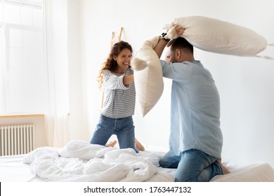 Happy young couple pillows fight and laughing on bed at bedroom. Smiling bearded man and woman family having fun and playing with cushions at home together.