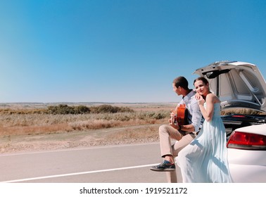 Happy young couple playing guitar and enjoying time together while sitting on open trunk of car parked on country road during summer trip