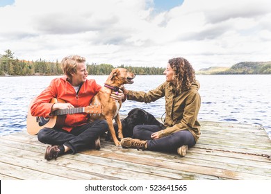 Happy young couple playing with their dog on a dock. The man is holding a guitar, they are wearing a rain jacket, love and nature concepts - Powered by Shutterstock