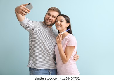 Happy young couple with pregnancy test taking selfie on color background
