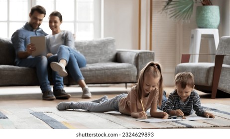 Happy young couple and preschooler children enjoying weekend at home. Kids rest on heating floor, playing and drawing, parents relaxing on couch, using tablet. Family leisure time in modern cozy house - Powered by Shutterstock