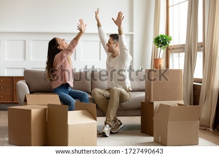 Happy young couple raising hands, sitting on couch in living room with cardboard boxes, celebrating moving day, family excited by relocation into first new house, buying real estate, mortgage