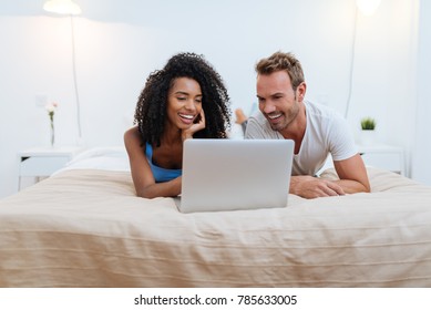 Happy Young Couple Relaxed At Home Lying Down In Bed On The Computer