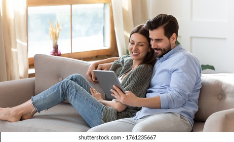 Happy Young Couple Relaxing On Cozy Couch, Using Computer Tablet Together, Smiling Beautiful Woman And Man Hugging, Looking At Device Screen, Watching Movie Or Video, Shopping Online, Browsing Apps