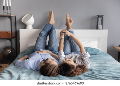 Happy young couple relaxing on bed together, using smartphone, looking at screen, man and woman enjoying lazy weekend in bedroom, shopping online, browsing mobile device apps, surfing internet - Powered by Shutterstock