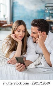 Happy Young Couple Relaxing On Bed At Home In Bathrobes, Using Mobile Phone, Taking Selfie Or Scrolling Social Media Together, Having Fun. Real People Lifestyle, Emotions. Date. 
