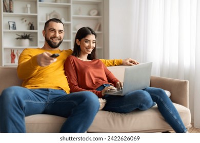 Happy Young Couple Resting On Couch At Home, Man Watching Tv And Woman Using Laptop, Cheerful Millennial Spouses Spending Time Together In Living Room, Enjoying Domestic Leisure, Closeup - Powered by Shutterstock