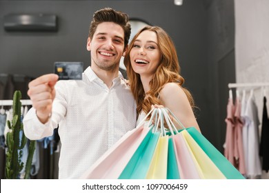 Happy Young Couple Shopping For Clothes Together At The Clothing Store, Woman Holding Shopping Bags And Man Showing Credit Card