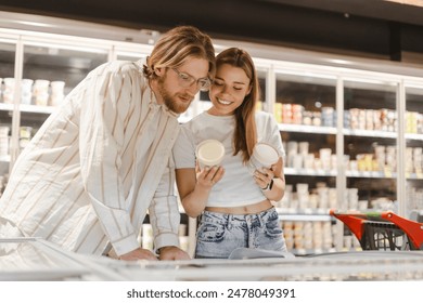 Happy young couple shopping for groceries, reading a product label while choosing frozen food from a supermarket fridge, doing grocery shopping together - Powered by Shutterstock