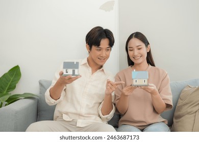 Happy young couple showing a small model house together, symbolizing home ownership, future investment or real estate concept, with a cozy living room interior in the background - Powered by Shutterstock