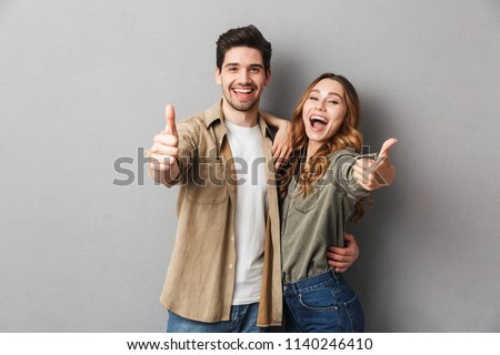 Happy young couple showing thumbs up and looking at camera isolated over gray background