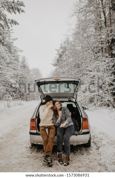 Happy Young\
couple is sitting in the back trunk of car on winter road and\
drinking hot tea from thermos. Winter\
time