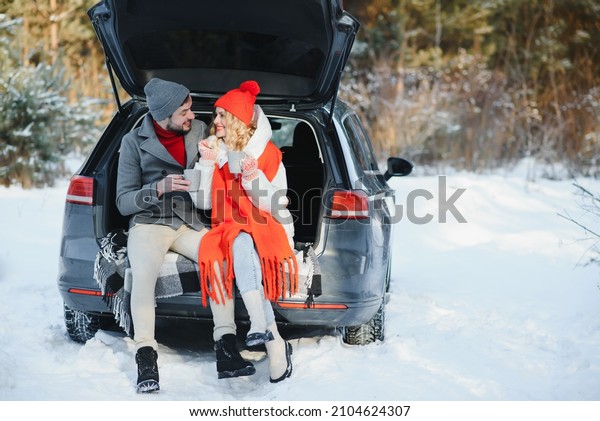 Happy\
Young couple is sitting in the back trunk of car in winter forest\
and drinking hot tea from thermos. Winter\
time