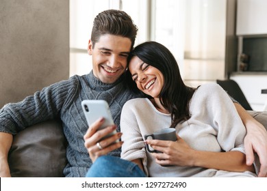 Happy Young Couple Sitting On A Couch At Home, Drinking Tea, Looking At Mobile Phone