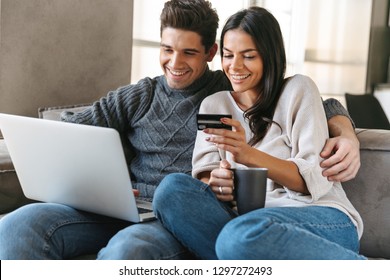 Happy young couple sitting on a couch at home, using laptop computer, showing credit card - Powered by Shutterstock