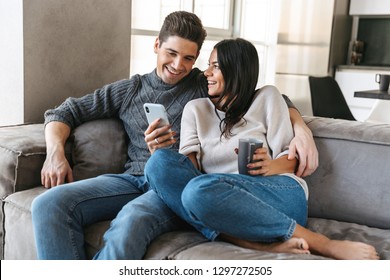 Happy Young Couple Sitting On A Couch At Home, Drinking Tea, Looking At Mobile Phone