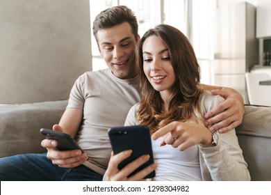 Happy Young Couple Sitting On A Couch At Home, Using Mobile Phone