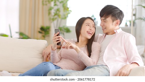 Happy Young Couple Sitting On  Couch And  Looking At Mobile Phone