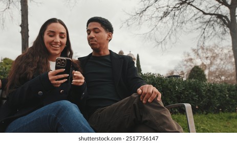 Happy young couple sitting on bench outdoors, sharing moment while looking at smartphone together - Powered by Shutterstock