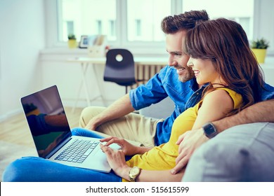 Happy Young Couple Sitting On Sofa At Home With Laptop