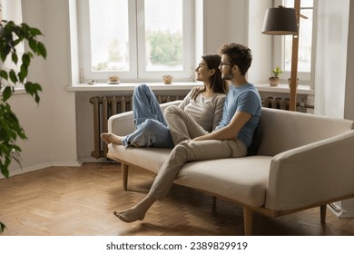 Happy young couple spend free time on sofa in living room looking out window, smiling, discuss plans and bright joint future, enjoy leisure time at home, daydreaming, resting at home. Family pastime - Powered by Shutterstock