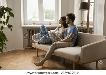 Happy young couple spend free time on sofa in living room looking out window, smiling, discuss plans and bright joint future, enjoy leisure time at home, daydreaming, resting at home. Family pastime