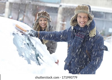 Happy Young Couple Standing In Snowfall, Brushing Off Snow From Car.