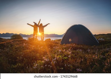 Happy young couple stands with hands up at sunset near tent  camping on a sunset background, traveling wanderlust in Norway. Lofoten Islands.
