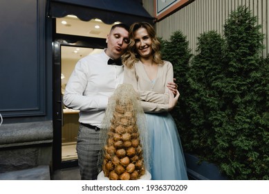 Happy Young Couple Stands Near The Birthday Cake. Unique Wedding Cake For Newlyweds. Husband And Wife Bite The Cake. Romantic Dessert