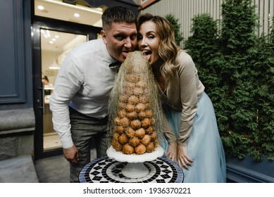 Happy Young Couple Stands Near The Birthday Cake. Unique Wedding Cake For Newlyweds. Husband And Wife Bite The Cake. Romantic Dessert