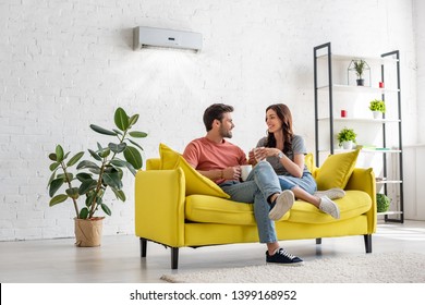 happy young couple talking and holding cups while sitting on yellow sofa under air conditioner at home - Powered by Shutterstock