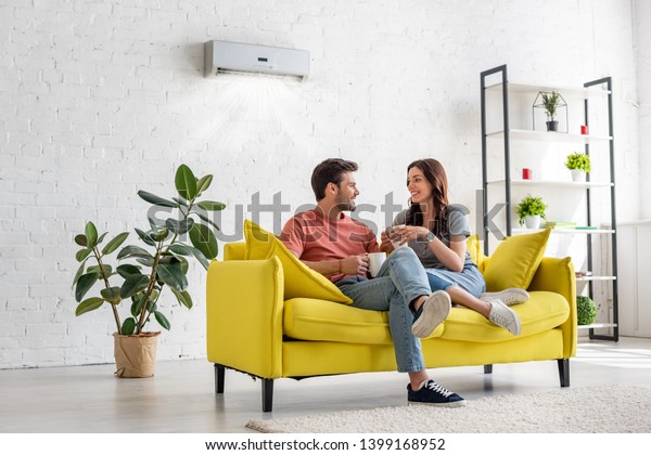 happy young couple talking\
and holding cups while sitting on yellow sofa under air conditioner\
at home