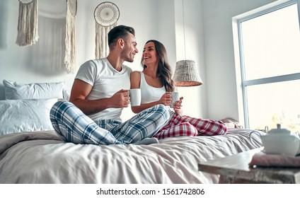 Happy Young Couple Talking And Laughing While Sitting On  The Bed And Drink Coffe In The Morning At Home.