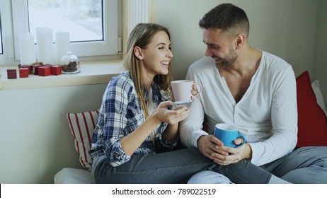 Happy young couple talking and laughing while sitting in on bed and drink coffe in the morning at home