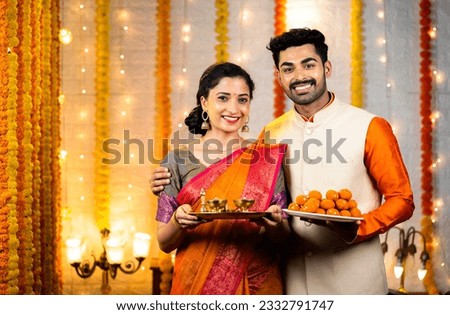 Happy young couple with traditional ethnic wear looking camera by holding sweets and pooja thali or plates - conept of festival celebration, rituals and cultural beliefs