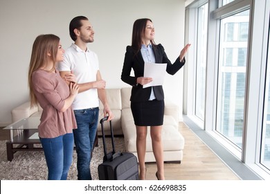 Happy Young Couple Travelers Moving In New Rented Own Apartment, Standing By The Window Enjoying View, Meeting With Female Real Estate Agent Showing Flat For Rent, Telling About Nearby Attractions 